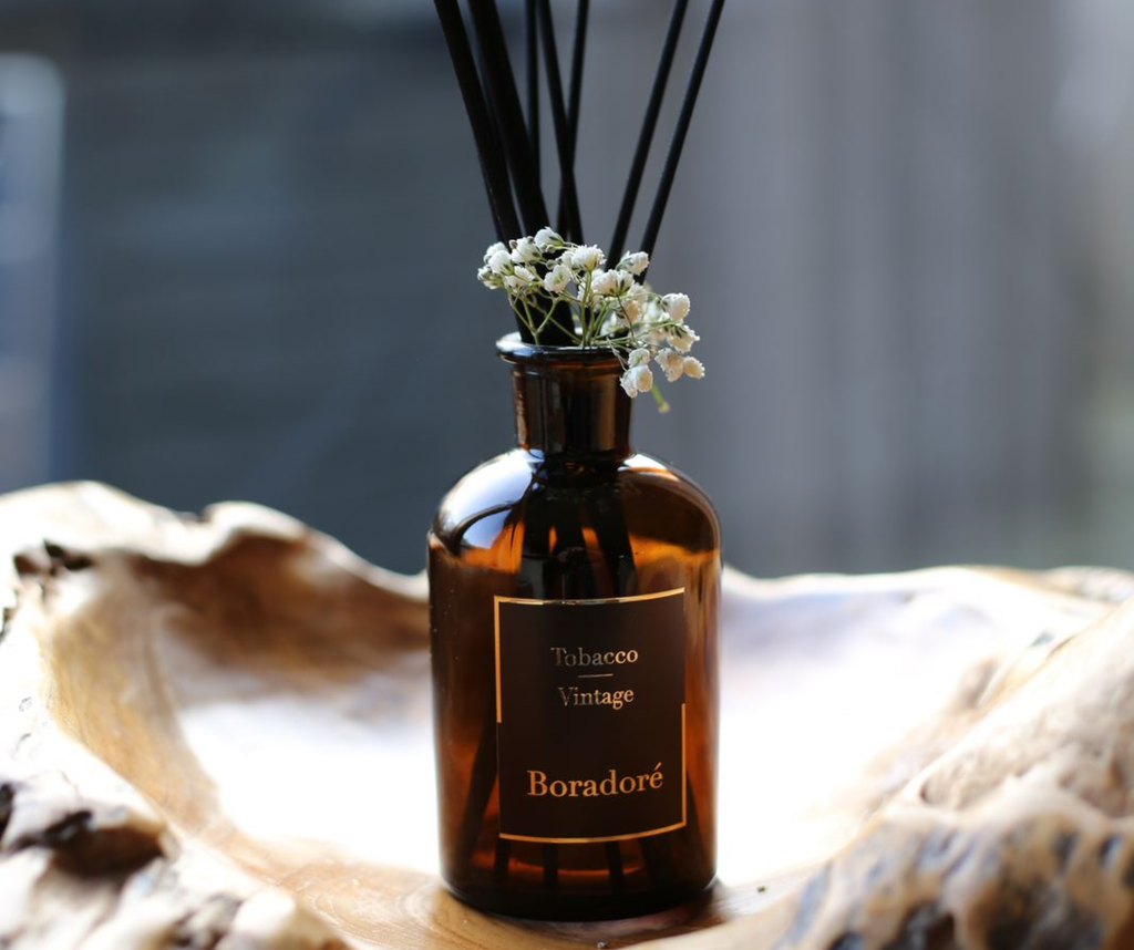 Boradoré Tobacco Vintage reed diffuser with black reeds and delicate white flowers in an amber glass bottle
